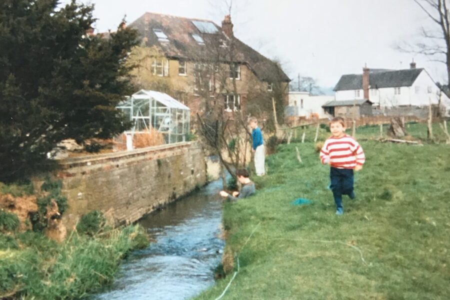 A photograph of a photograph of Michael as a child, in a white and red jumper running beside a stream in a field with two boys behind him and some houses in the distance.