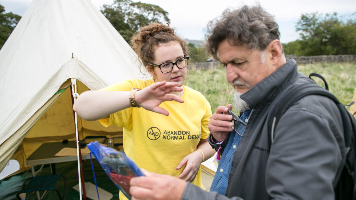 AND Festival volunteer helping someone use an iPad. They are outside and next to a yurt.